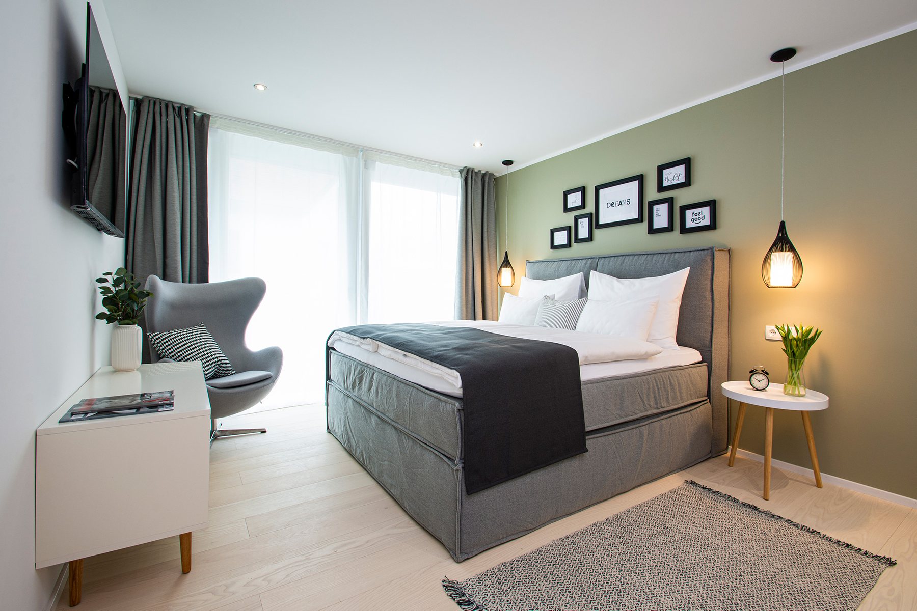Hotelzimmer mit Doppelbett, grüner Wand mit Bilderrahmen und Pendelleuchten